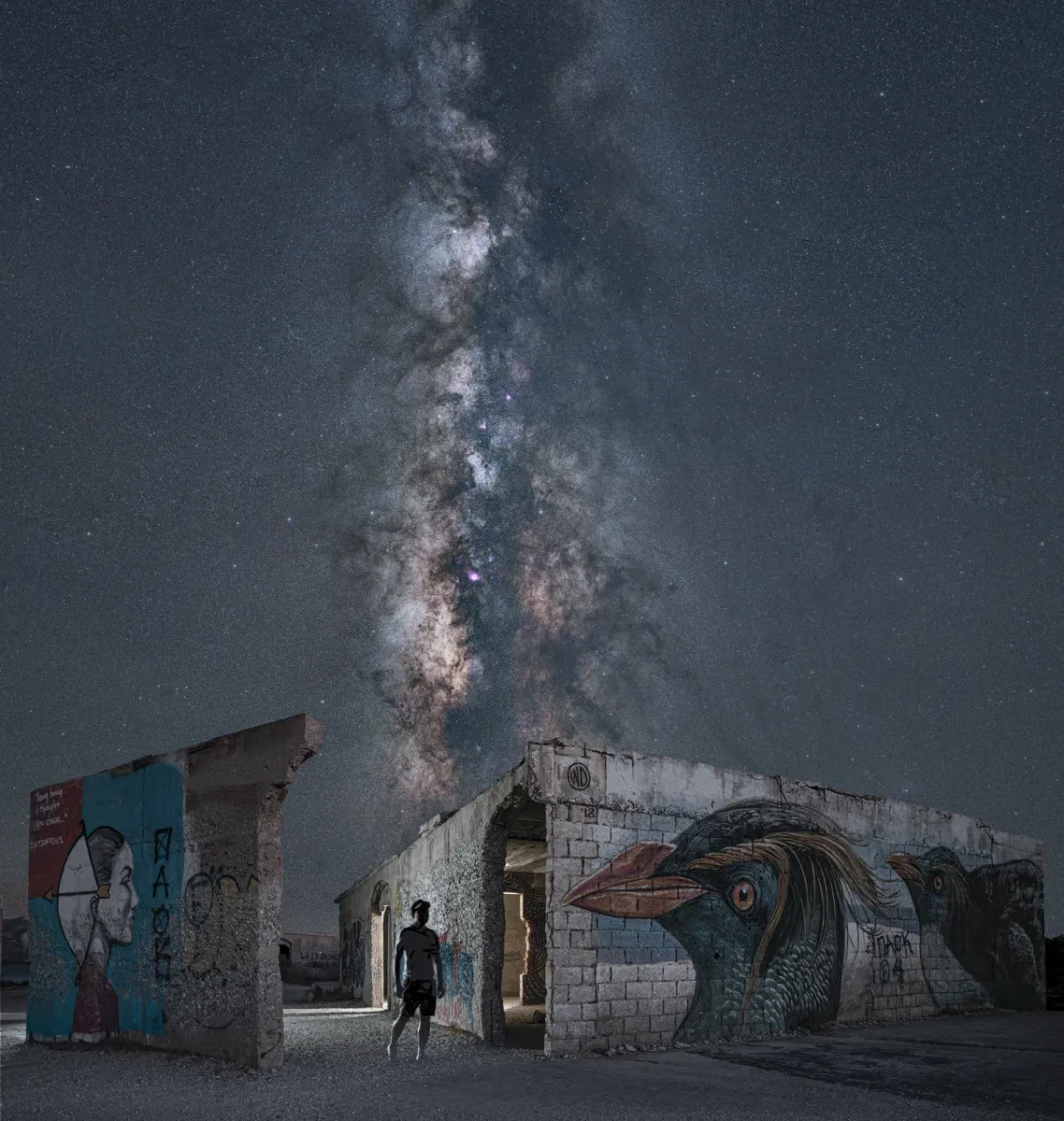 A night sky photograph of the Milky Way taken amongst ruined buildings. The eerie scene features graffiti on crumbling walls, with the figure of a man in shadow standing by a painting of a bird. A stream of stars bisects the sky above them